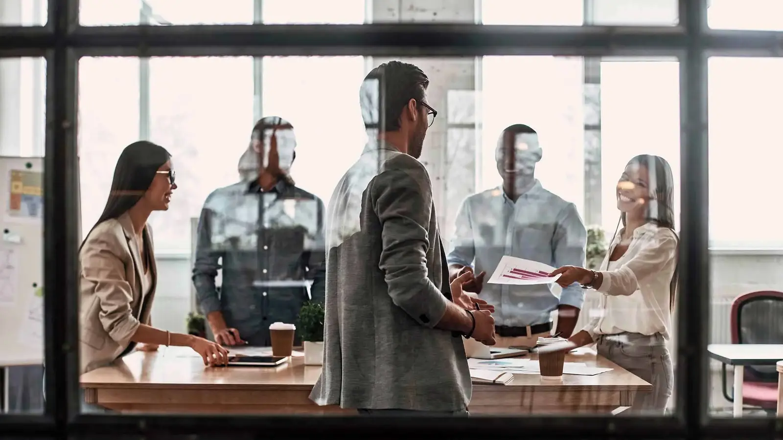  Hinter einer großen Glaswand versammeln sich mehrere Büroangestellte zu einem Meeting und tauschen Dokumente miteinander aus. 