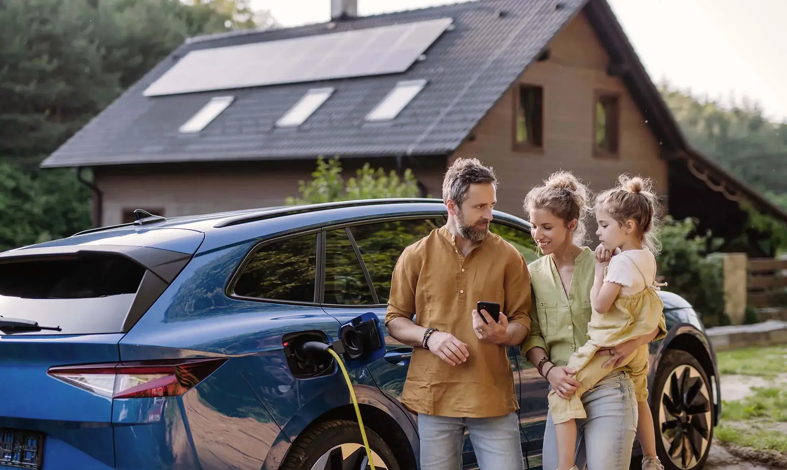 Eine Familie, ein Vater, die Mutter und ein Kind, stehen vor ihrem E-Auto, welches in der Nähe des Hauses geparkt ist. Das E-Auto wird geladen und der Vater zeigt der Mutter etwas auf seinem Handy. 